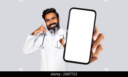 Un medico indiano che indossa un cappotto bianco con uno stetoscopio intorno al collo sorride e fa un gesto chiamato con le dita tenendo in mano uno smartphone W Foto Stock