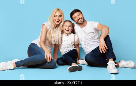 Una famiglia felice di tre persone, una madre, un padre e una figlia, si siedono insieme su uno sfondo azzurro. La famiglia indossa abiti casual e sorride Foto Stock
