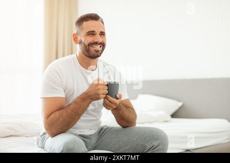 Un uomo siede su un letto, indossa una t-shirt bianca e pantaloni in felpa grigi, tiene in mano una tazza scura, sorride mentre guarda di lato. L'impostazione è Foto Stock