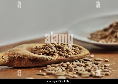Cucchiaio di legno con porzione di semi di girasole sgusciati e semi sparsi sul tavolo fuori fuoco. Foto Stock