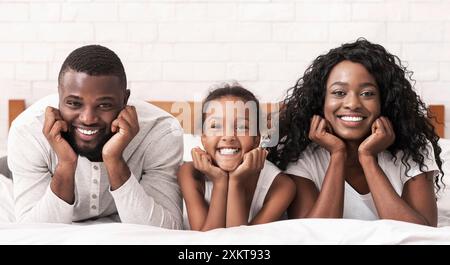 Ritratto di una famiglia nera felice sdraiata sul letto, con le mani in mano e sorridente alla fotocamera, copia spazio Foto Stock