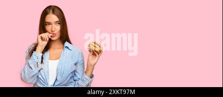 Amante del cibo spazzatura. Hungry Girl Holding Burger Licking Fingers in piedi su sfondo giallo Studio. Foto Stock