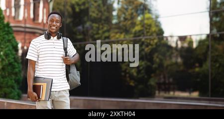 Felice studente afro-americano sorridente con zaino e libri di lavoro sul background universitario, guardando la macchina fotografica e sorridendo Foto Stock