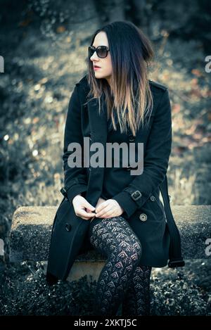 Una giovane donna che indossa un cappotto nero e calze alte sulla coscia si siede su un tronco intempestivo in un ambiente forestale. I suoi capelli scuri le incorniciano il viso e lei indossa Foto Stock