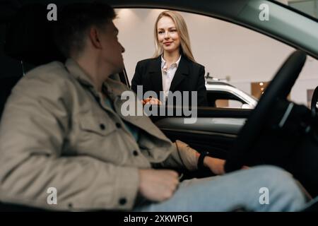 Guarda dall'interno del veicolo all'accogliente manager bionda che parla con un cliente maschio seduto in auto nello showroom che sceglie l'automobile. Concetto di Foto Stock