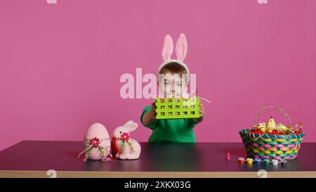 Dolce ragazzino che si occupa di allestimenti colorati per le festività natalizie di Pasqua, mettendo uova dipinte in un cestino artigianale. Un bambino entusiasta e gioioso che utilizza materiali per creare decorazioni. Telecamera B.. Foto Stock