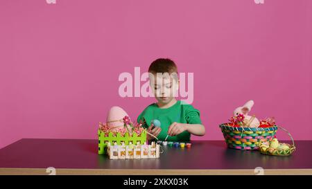 Gioioso bambino che dipinge uova per le festività natalizie di pasqua in studio, utilizzando acquerelli e forniture d'arte. Sorridente colorazione in età prescolare ornamenti festivi in preparazione per la domenica. Telecamera B.. Foto Stock