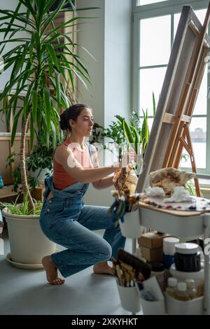 L'artista femminile interessata applica pittura a olio con pennello al disegno su tela su cavalletto in studio d'arte. Foto Stock