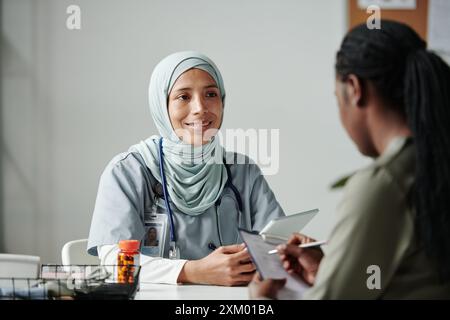 Giovane donna sorridente medico in hijab utilizzando tablet e consultare paziente afroamericano compilando il modulo medico durante la visita del medico Foto Stock