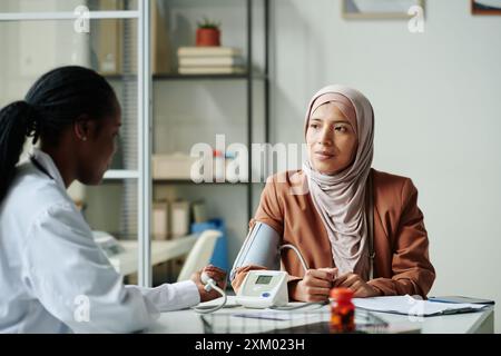 Giovane paziente musulmana in hijab e abbigliamento casual che guarda un medico afroamericano che misura la pressione sanguigna della donna Foto Stock