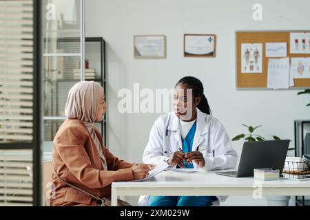 Giovane medico afroamericano in camice da laboratorio che parla con paziente musulmano in hijab e abbigliamento casual durante la consultazione medica Foto Stock