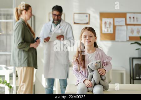 Una bambina sorridente e simpatica con un morbido coniglio grigio in mano che guarda la macchina fotografica mentre si siede contro la madre e si consulta con un pediatra Foto Stock