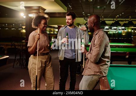 Gruppo di amici interculturali felici in abbigliamento casual che bevono birra e chiacchierano dopo aver giocato a biliardo mentre si trovava in sala giochi Foto Stock