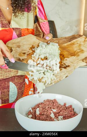 Primo piano di una donna che spruzza cipolle in un recipiente di carne macinata fresca, verdure ed erbe aromatiche in cucina. Foto Stock