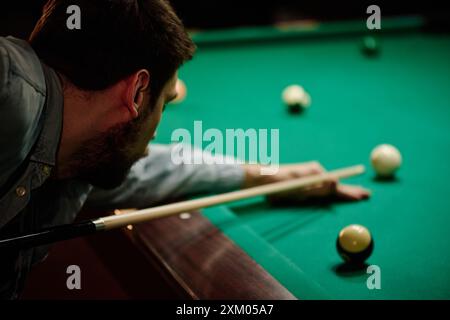 Concentrati su un giovane giocatore di biliardo maschile in abbigliamento casual che tiene in mano il segno mentre si piega sul tavolo verde e gioca nella sala giochi Foto Stock