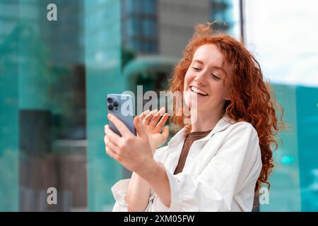 Ritratto di una giovane donna rossa in camicia bianca che parla e usa sul suo cellulare in una città moderna e urbana Foto Stock