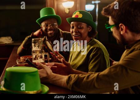 Giovane donna afroamericana in cappello verde seduta accanto al bancone del bar tra due ragazzi interculturali e che parla con loro Foto Stock