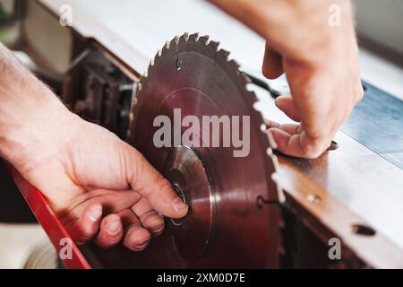 Abile falegname che sostituisce la lama di una sega da tavolo nella sua officina, si prepara alla lavorazione del legno con precisione e attenzione alla sicurezza. La segatura riempie l'aria come h Foto Stock