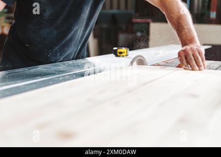 Carpentiere esperto che taglia il legno con sega circolare in un'officina affollata e che ruota la segatura. Primo piano mostra precisione, competenza e dedizione all'artigianato. Han Foto Stock