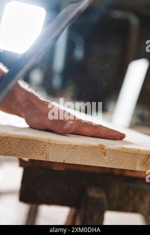 Un carpentiere esperto ispeziona la tavola di legno in officina, presentando una superficie liscia. La polvere di segatura crea lo sfondo mentre crea mobili con precisione, aggiungendo Foto Stock