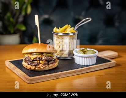 Doppio hamburger di manzo con formaggio, sottaceti e salsa si trova su un'ardesia nera con patatine fritte e sottaceti Foto Stock