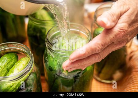 La mano di una persona anziana versa con cura acqua bollente in vasetti riempiti di cetrioli freschi, mostrando il metodo tradizionale di decapaggio del sale. Idea Foto Stock