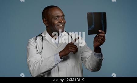 Pensivo sorridente uomo afroamericano maschio radiologo medico chirurgo traumatologo che esamina radiografia in studio blu background check risultati scansione Foto Stock