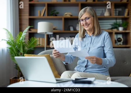 Donna anziana seduta sul divano che esamina i documenti mentre lavora da casa con un laptop. Si concentra e si adatta a contenuti in un ambiente accogliente, impegnato in attività di lavoro a distanza o personali Foto Stock