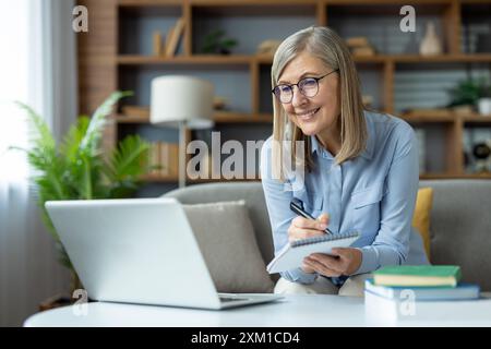 Donna matura che studia online con un portatile, scrive appunti, sorride mentre impara a casa. Apprendimento permanente, istruzione e studio a distanza Foto Stock