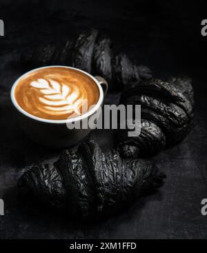 Croissant neri gourmet con una tazza di cappuccino su sfondo nero. Vista dall'alto verticale. Foto Stock