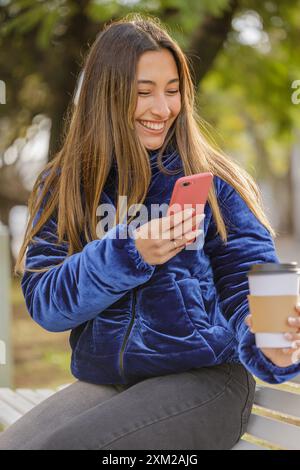 Una ragazza latina seduta su una panchina del parco pubblico scatta una foto della sua tazza da caffè usa e getta. Foto Stock