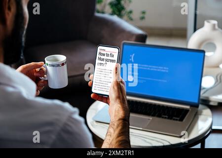 Uomo con telefono cellulare alla ricerca di informazioni per correggere la schermata blu della morte o BSOD del suo portatile. Errore sistema operativo difettoso. Foto Stock