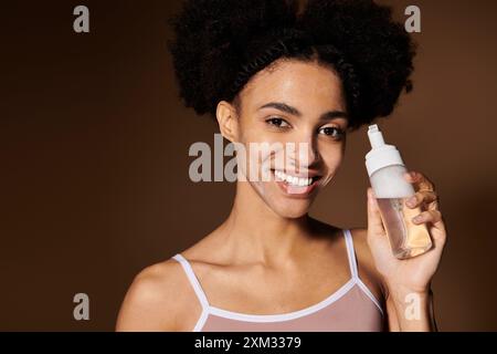 Una giovane donna afroamericana sorride brillantemente, tenendo in mano una bottiglia di schiuma per il viso. Foto Stock