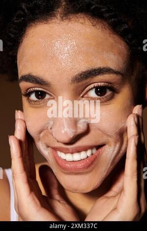 Una giovane donna afroamericana sorride mentre pulisce il viso con un detergente schiumoso. Foto Stock