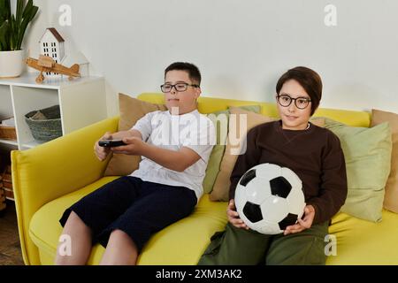 Due ragazzi, uno con una palla da calcio, rilassati su un divano giallo in un ambiente domestico. Foto Stock