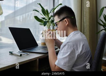 Un ragazzo con la sindrome di Down siede a una scrivania in casa sua, concentrandosi su un compito. Foto Stock