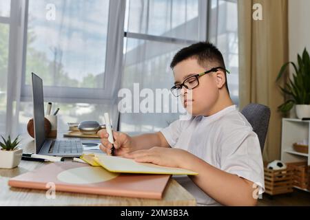 Un ragazzo con la sindrome di Down si concentra sui suoi studi a casa, utilizzando un notebook e un notebook. Foto Stock