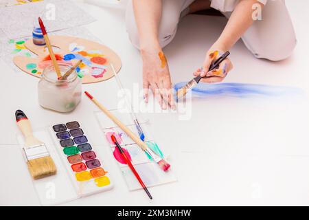 Persona che dipinge con acquerelli sul pavimento, mani con vernice, pennelli, tavolozza e un barattolo. Spazio di lavoro creativo con strumenti artistici Foto Stock