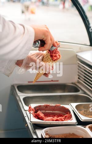 Cono per waffle con delizioso gelato alla fragola in mano. Il venditore mette una palla di gelato in un cono per waffle con un cucchiaio o un cucchiaio Foto Stock