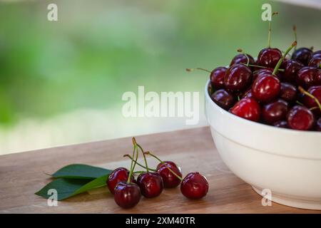 Ciliegie fresche con steli e foglie su tavola di legno, ciotola di ciliegie sullo sfondo con ombreggiatura a foglia. Foto Stock