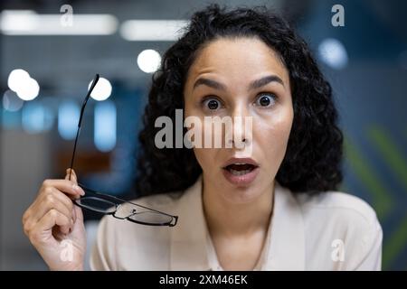 Un impiegato ha sorpreso gli occhiali in mano con un'espressione sconvolgente in un ambiente d'ufficio moderno. Donna in abbigliamento professionale che rappresenta l'emozione. Concetto di shock, sorpresa e notizie inaspettate funzionano. Foto Stock