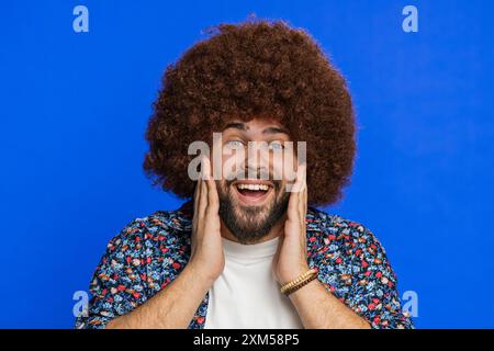 Oh mio Dio, Wow. Vincitore sorridente con parrucca Afro che solleva le mani di sorpresa guardando la telecamera con grandi occhi scioccato dalla vittoria che celebra il successo buone notizie. Ragazzo su sfondo blu Foto Stock