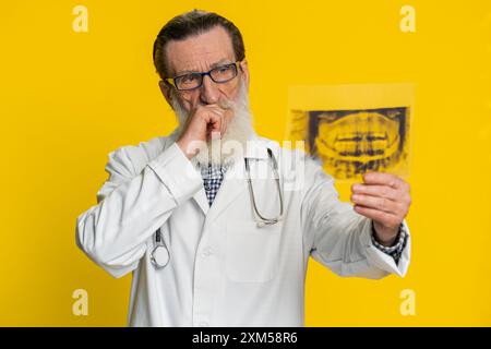 Il medico anziano maturo ortodontista esamina una foto radiografica panoramica dei denti della mascella. Modello 3D della bocca del paziente, scansione RM. Odontoiatria orale. Nonno di Stomatologia su sfondo giallo Foto Stock