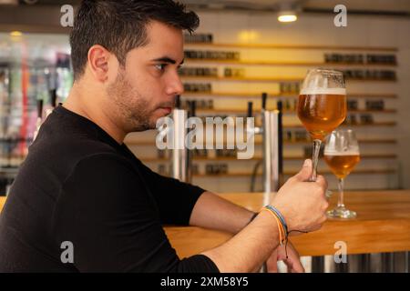 Un giovane latino si gode una birra artigianale in un pub. Osserva attentamente il colore e il luppolo nella lager che sta assaggiando. Foto Stock