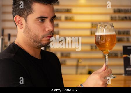 Un giovane latino si gode una birra artigianale in un pub. Osserva attentamente il colore e il luppolo nella lager che sta assaggiando. Foto Stock