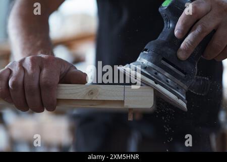 Carpenter sta levigando un pezzo di legno nella sua officina, utilizzando una levigatrice orbitale. Indossa una maglietta nera e si trova di fronte a un banco da lavoro Foto Stock