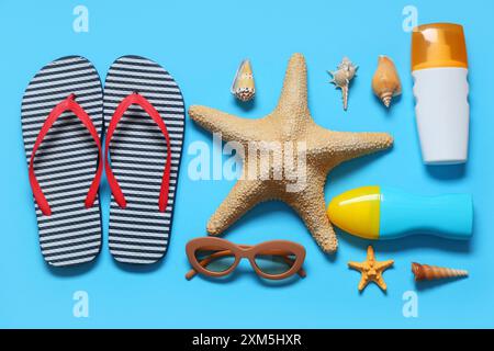 Eleganti infradito a righe con conchiglie, occhiali da sole, stelle marine e bottiglie di crema solare su sfondo blu Foto Stock