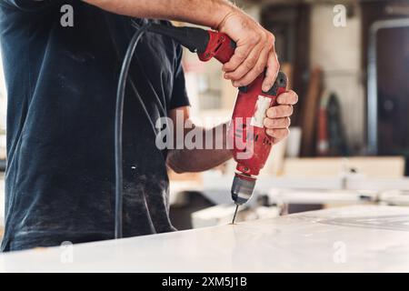 Carpenter sta realizzando un foro su una tavola di legno bianca utilizzando un trapano elettrico in un'officina. Il falegname indossa una maglietta nera e utilizza una corda rossa Foto Stock