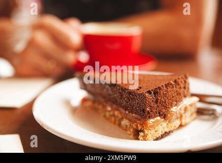 Appetitosa fetta di torta al cioccolato e uomo con una tazza di caffè rosso su sfondo sfocato Foto Stock