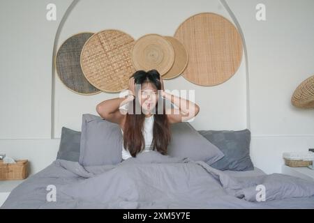 La donna asiatica che si sveglia la mattina si sente stressata e ha mal di testa dopo un sonno profondo, ma si sente ancora stanca e sonnolenta stare seduta sul letto nel Foto Stock